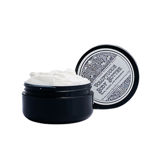 A jar of body butter with its lid off, showing a creamy white product inside. The jar is placed on a surface with a decorative lid.